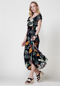 Vestido maxi negro con estampado floral, que presenta un escote en V, mangas cortas y un dobladillo fluido. La tela tiene patrones de hojas coloridos. Combinado con sandalias de cuña.