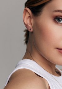 Czarne kolczyki wkrętki noszone w lewym uchu, o gładkiej powierzchni i minimalistycznym designie, uzupełniają jasny bezrękawnik.