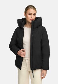 Vrouw in een zwart gewatteerd jack met een grote capuchon, een lichtbeige geribbelde top en crèmekleurige broek tegen een effen lichte achtergrond.