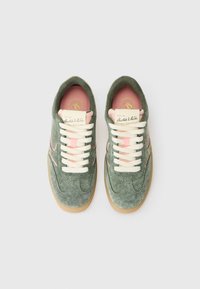 Une paire de baskets en daim vert avec des lacets blancs, des accents roses et des semelles en caoutchouc, vue d'en haut sur un fond blanc.