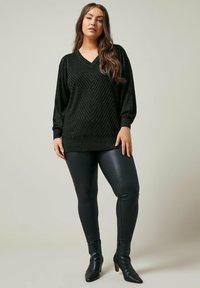 Maglione nero a trama con scollatura a V e motivo a zig-zag, abbinato a leggings neri lucidi e stivaletti a punta alla caviglia.