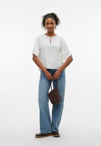 Kvinde i hvid kortærmet bluse og blå jeans, der holder en brun ruskinds håndtaske, stående mod en ensfarvet hvid baggrund.