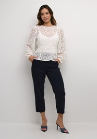 Hvid blonde top med lange ærmer og rund halsudskæring, parret med marineblå bukser og blå åbne hæle. Blonden har indviklede blomster mønstre.