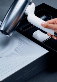 Hand hält weißen Aufsatz nahe an silbernem professionellem Haartrockner mit dualem Luftstrom, der auf einer strukturierten silbernen Box und einem schwarzen Tablett liegt.