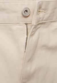 Close-up van beige denim broek met een metalen knoop waarop "MNG DENIM" staat en een gedeeltelijk zichtbare zilveren ritssluiting.