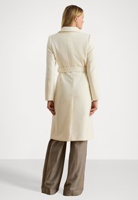 Kremkrāsas vilnas mēteli ar jostu viduklī, ar izgriezumu krūšu daļā un garām piedurknēm. Veidots ar izšuvumiem, lai nodrošinātu pielāgotu piegriezumu. Nēsāts virs brūnām plašām biksēm.