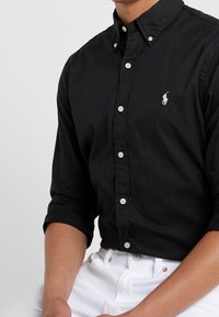 Czarna koszula zapinana na guziki, wykonana z miękkiego materiału, z kołnierzykiem, długimi rękawami podwiniętymi do połowy ramienia oraz małym białym logo na piersi.