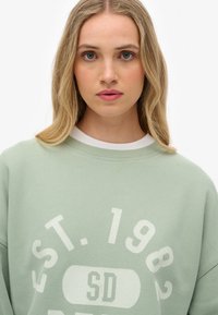 Sudadera verde menta con cuello redondo blanco, que presenta un texto sobredimensionado "EST. 1982 SD" y una textura suave. Diseño simple y ajuste casual.
