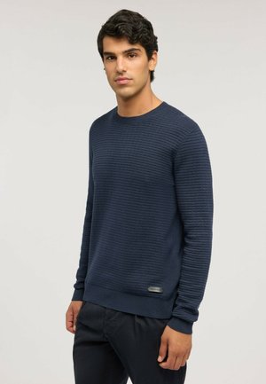 Giovane uomo con capelli ricci scuri indossa un maglione blu navy a maniche lunghe con texture e pantaloni scuri, in piedi contro uno sfondo chiaro e uniforme.
