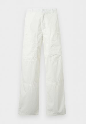 Νέος άνδρας στέκεται με τα χέρια στις τσέπες, φοράει γκρι σακάκι, μαύρο πουκάμισο, λευκό φαρδύ παντελόνι και μαύρα σανδάλια σε μονόχρωμο φόντο.