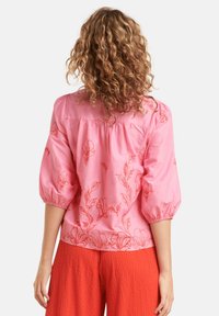 Rosa Bluse mit floraler Stickerei, Puffärmeln und gerafftem Rücken. Getragen mit strukturierten roten Hosen.
