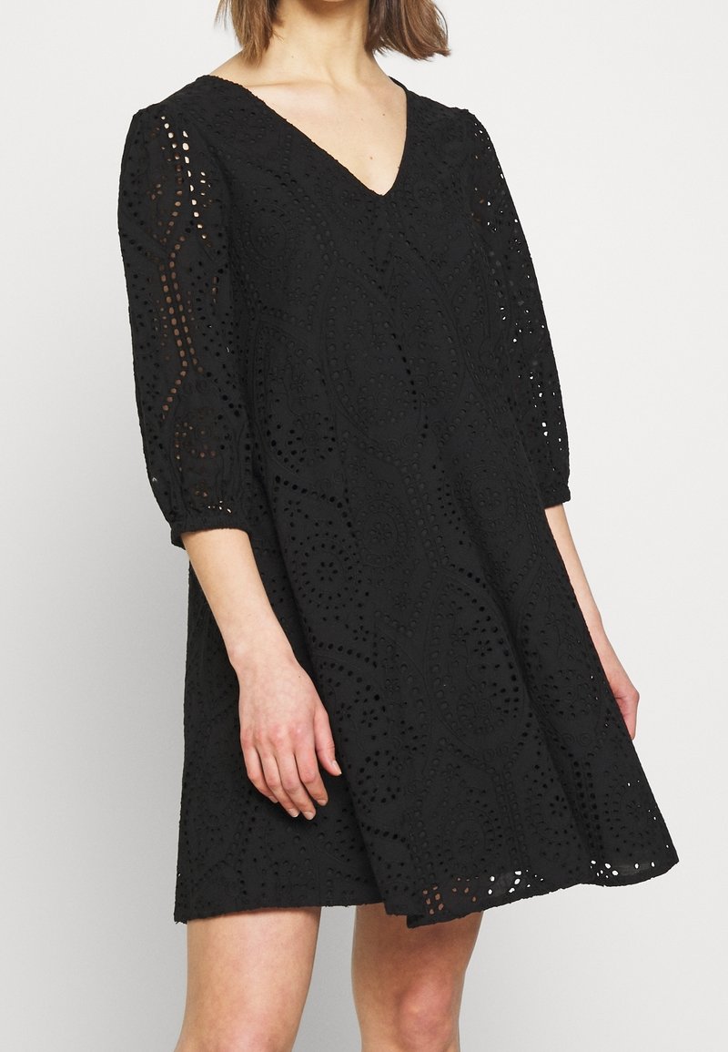 Musta mekko V-pääntiellä ja kolme neljäsosaa hihoilla, jossa on leikattuja kuvioita ja teksturoitu pinta, levenee helman kohdalla.