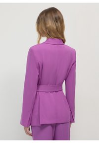 Dopasowana marynarka w jasnym purpurowym kolorze, wykonana z gładkiego materiału. Posiada pasek w talii, długie rękawy oraz dopasowany krój z detalem wentylacyjnym z tyłu.