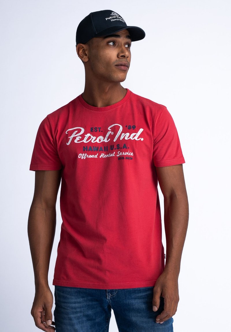Red cotton t-shirt with round neck, short sleeves, and front text print. Worn with blue jeans. Dark cap with logo on a model.