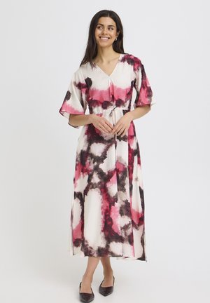 Femme souriante, vêtue d'une longue robe ample avec des motifs abstraits roses, noirs et blancs, debout les mains jointes devant elle.