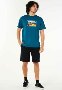 Teal kortærmet t-shirt med et retro-inspireret graphic og sorte shorts. Båret med sorte sneakers. Glat stof og afslappet pasform.