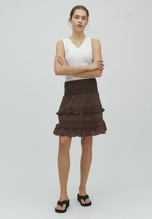 Młoda kobieta z założonymi rękami, ubrana w biały bezrękawnik, brązową warstwową spódnicę i czarne sandały na platformie, stojąca na tle jednolitego jasnego tła.