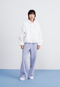 Bijela oversized jakna s kragnom, uparena s plavim i bijelim prugastim širokim hlačama. Bijele tenisice upotpunjuju look na jednoličnoj pozadini.