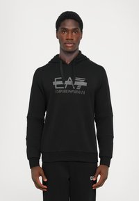 Črna kapuca z logotipom spredaj, ki prikazuje "EA" in "EMPORIO ARMANI." Narejena iz mehkega materiala, z sproščenim krojem in vrvico na kapuci.