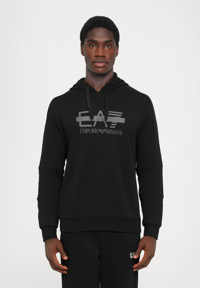 Črna kapuca z logotipom spredaj, ki prikazuje "EA" in "EMPORIO ARMANI." Narejena iz mehkega materiala, z sproščenim krojem in vrvico na kapuci.