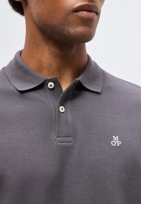 Κοντινό πλάνο ενός ατόμου που φοράει γκρι πόλο πουκάμισο με δύο λευκά κουμπιά κουμπωμένα και μικρό λευκό λογότυπο "MOP" στο στήθος.