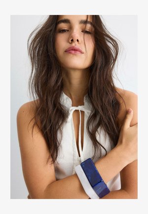 Jeune femme aux longs cheveux bruns ondulés porte un haut blanc sans manches et deux gros bracelets rectangulaires, un bleu et un blanc, à son bras.