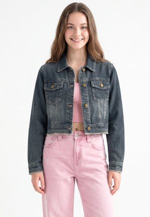 Giovane donna sorridente che indossa una giacca corta in denim scuro, un crop top rosa e jeans rosa chiaro a vita alta su sfondo bianco.