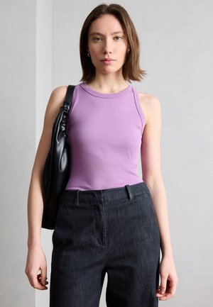 Γυναίκα με καστανά μαλλιά μέχρι τους ώμους, φορώντας αμάνικη λεβαντί μπλούζα και σκούρο παντελόνι ψηλής μέσης, κρατώντας μια μαύρη τσάντα ώμου.