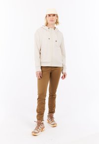 Khaki rits hoodie met trekkoord, beige fitted broek en lichtbruine wandelschoenen met veters en gestructureerde zolen. Pet in bijpassend beige.