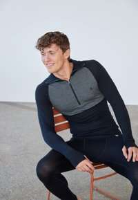 Langärmeliges Sportshirt mit einem halben Reißverschluss, das einen grauen oberen Teil und marineblaue Ärmel aufweist, kombiniert mit eng anliegenden dunklen Hosen.