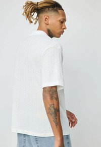 Chemise blanche à manches courtes avec un motif texturé, un col et une coupe décontractée. Les tatouages du modèle sont visibles sur le bras, vus de derrière.