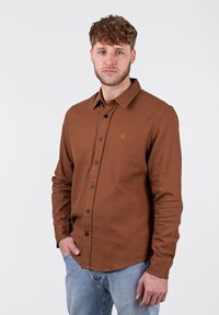 Camisa marrón de botones con cuello, botones negros y un logo sutil en el pecho; combinada con jeans de color azul claro.
