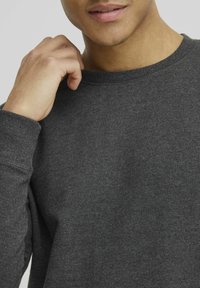 Dark gray sweatshirt in cotton blend, featuring a ribbed crew neck and textured fabric. Close-up shows relaxed fit and sleeve details.