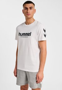 Vit bomull t-shirt med svart "hummel"-logotyp och text, korta ärmar med svarta chevronmönster, i kombination med grå shorts.