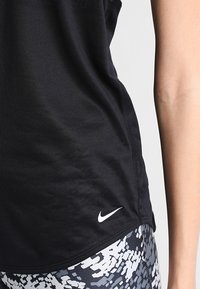 Canottiera nera atletica realizzata in tessuto liscio, con un piccolo logo Nike bianco sul lato. Abbinata a leggings grigi a fantasia.