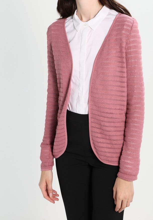 Femme portant un cardigan ouvert texturé rose poussière sur une chemise blanche à col et un pantalon noir sur fond uni.