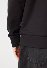 Sweatshirt preto com punhos e bainha em malha, exibindo uma textura suave. Pequeno etiqueta na lateral com o texto "BOSS". Usado com jeans cinzentos.