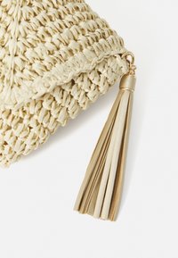 Sac en paille tressée de couleur crème avec une forme triangulaire texturée, doté d'un accent en pompon doré et d'une attache en métal.