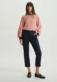 Chemise à manches longues rayée rouge et crème avec des accents noirs, associée à un pantalon court bleu marine foncé et des mocassins marron. Design simple.