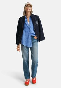 Marineblazer met witte stiksels en een embleem, lichtblauw overhemd, ontspannen blauwe jeans en rode sneakers met witte veters.