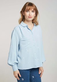 Chemise bleu clair à boutons avec un tissu texturé, poche poitrine unique et accents de broderie florale blanche. Manches longues avec un poignet froncé.