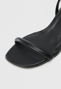 Black sandal with a sleek design, featuring a thin strap over the toes and a textured footbed. The material appears smooth and synthetic.