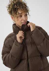 Hnedá páperová bunda s vysokým golierom, zipsovým zapínaním a prešitými sekciami. Hladká textúra s jemným leskom.