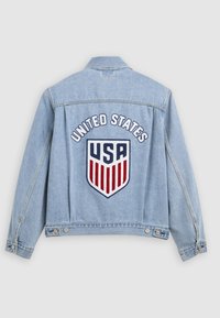 Ανοιχτό μπλε μπουφάν τζιν με το κείμενο "United States" και το λογότυπο με τη ζωστήρα της ΗΠΑ σε κόκκινο, άσπρο και μπλε στην πλάτη.