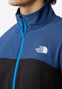 Blue and black zip-up jacket with high collar, featuring a white logo on the chest and contrasting blue zipper detail. Lightweight fabric.