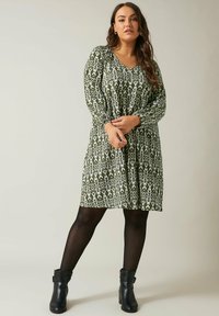Grünes gemustertes Kleid mit V-Ausschnitt, langen Ärmeln und einer A-Linien-Silhouette. Präsentiert weiße abstrakte Designs. Getragen mit schwarzen Knöchelstiefeln.