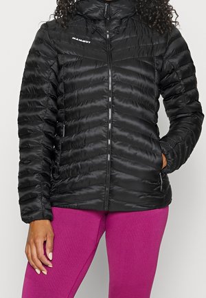 Žena oblečená v černé prošívané bundě Mammut s kapsami na zip a kapucí, v kombinaci s jasně růžovými texturovanými legínami.