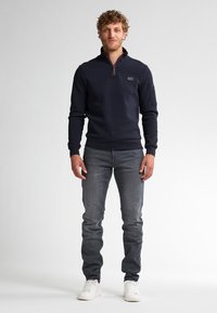 Marineblauwe zip-up sweatshirt met ribbelboorden bij de mouwen en onderkant, voorzien van een borstlogo. Gekoesterd met goed zittende grijze jeans en witte sneakers.
