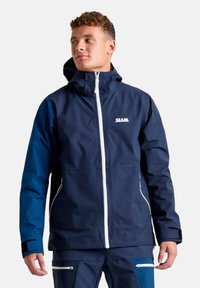 Giacca impermeabile blu navy con colletto alto, zip bianca e logo. Presenta maniche blu a contrasto e tasche con zip.