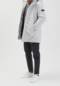 Hellgrauer Parka mit Kapuze, Fronttaschen und schwarzem Akzentpatch; kombiniert mit dunklen Hosen und weißen Sneakers. Glattes, isoliertes Material.